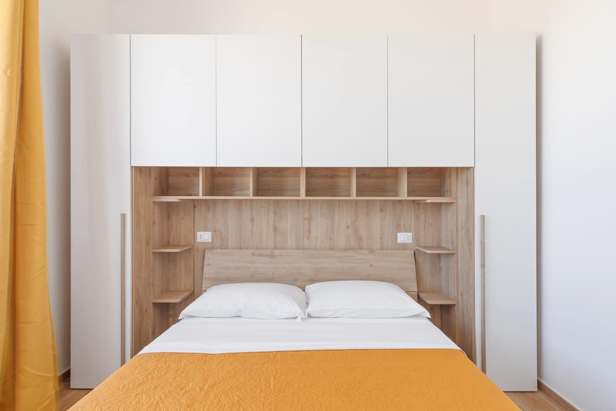 A bedroom with a bed, white headboard, wooden shelves, and a yellow bedspread.