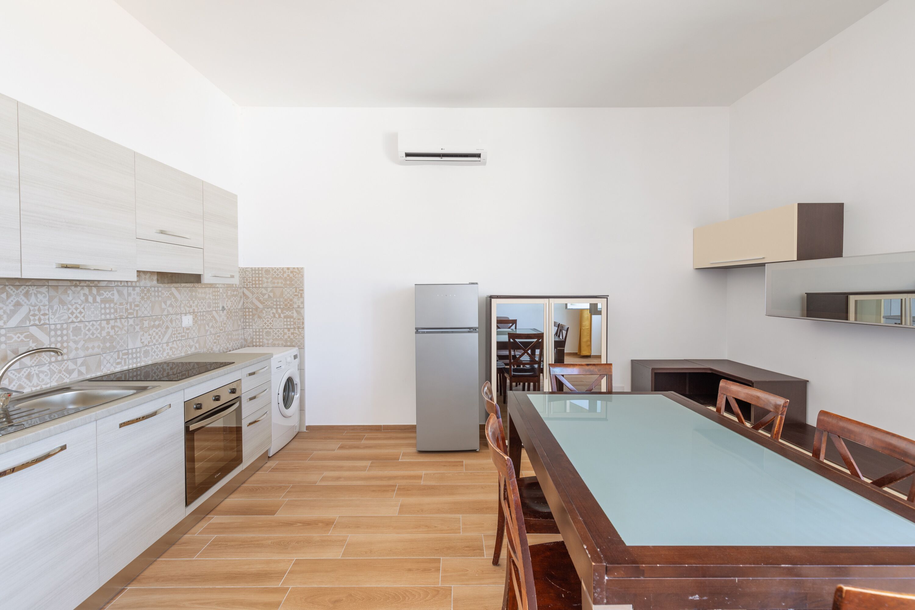 A modern kitchen with white cabinets, a built-in oven, a washing machine, and a dining area with a glass table and wooden chairs.
