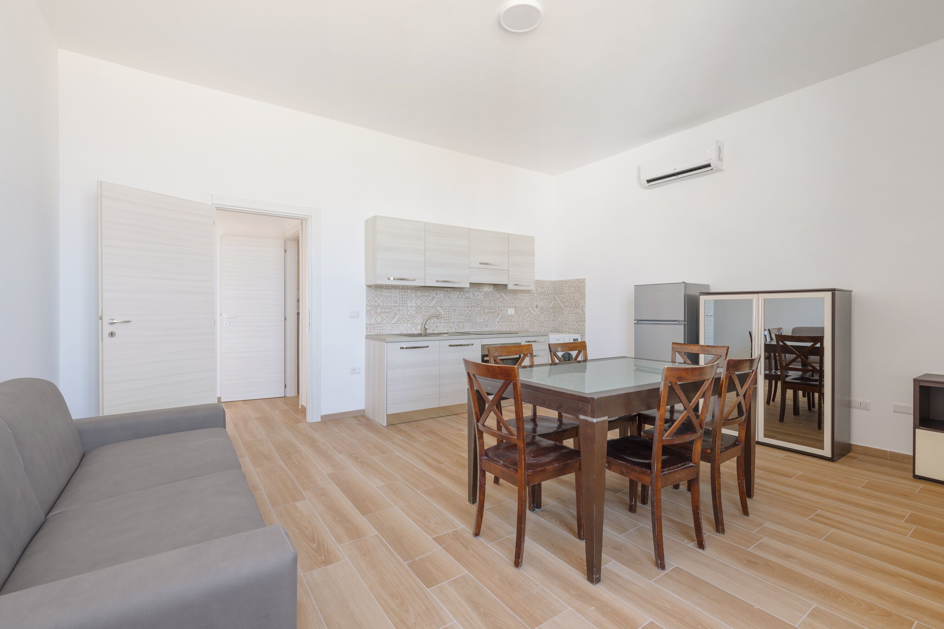 A modern kitchen with white cabinets, a dining table with wooden chairs, and a grey sofa in a bright room with wooden flooring.