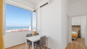 A room with a dining table and chairs, a bed with a yellow blanket, and a view of the beach and ocean through a large window. - Residence Torvaianica (Pomezia)