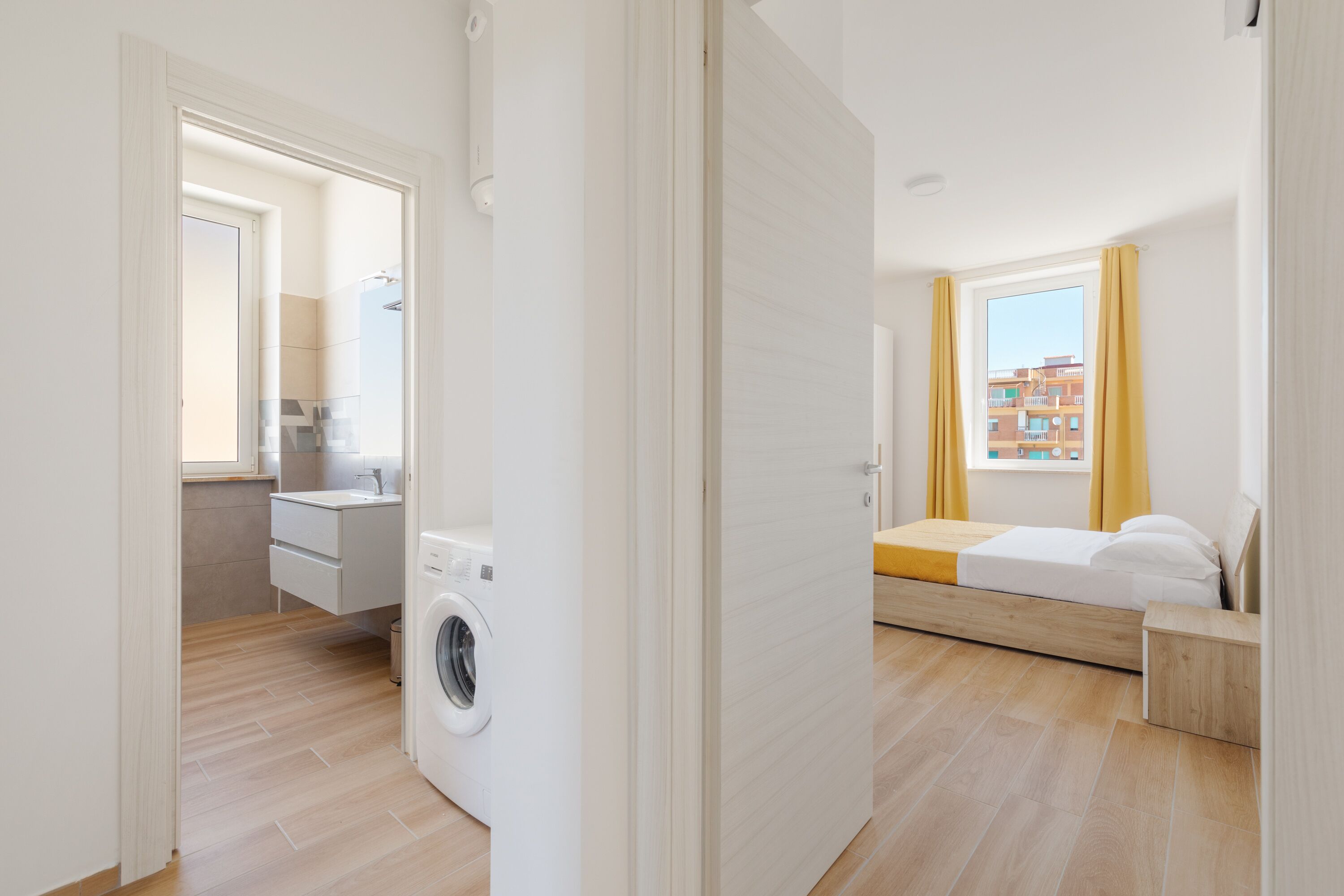 En moderne lejlighed med vaskefaciliteter, et lille tekøkken, en seng og et vindue med gardiner.