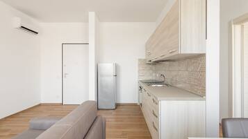 Una cocina moderna con horno integrado, refrigerador y lavamanos. En la zona de estar, hay una mesa de comedor de madera y un sofá gris.