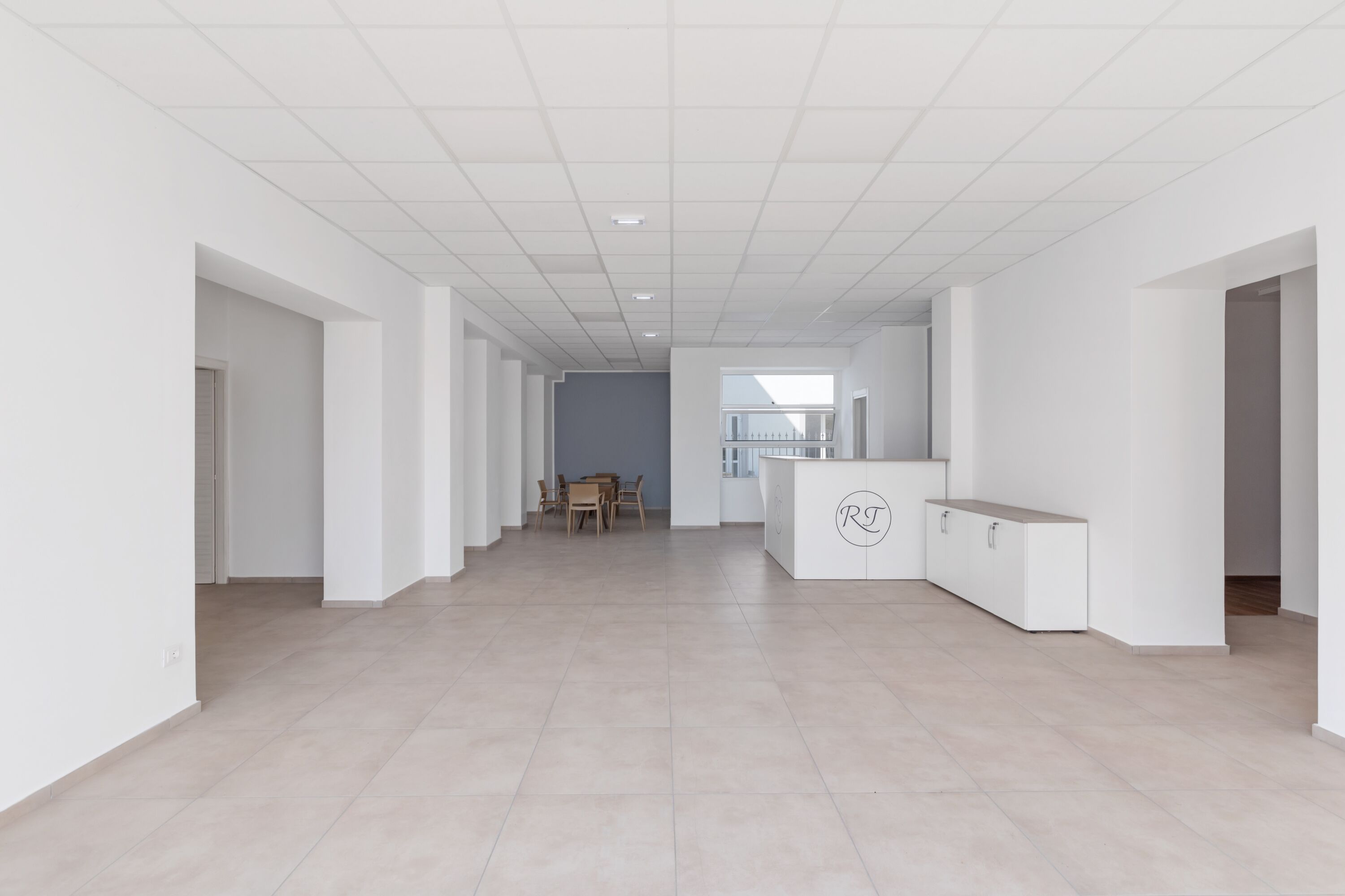 A spacious, modern hallway with a neutral color palette, featuring a dining area with a table and chairs, a reception desk, and a view of a balcony.