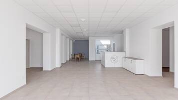 A spacious, modern hallway with a neutral color palette, featuring a dining area with a table and chairs, a reception desk, and a view of a balcony.