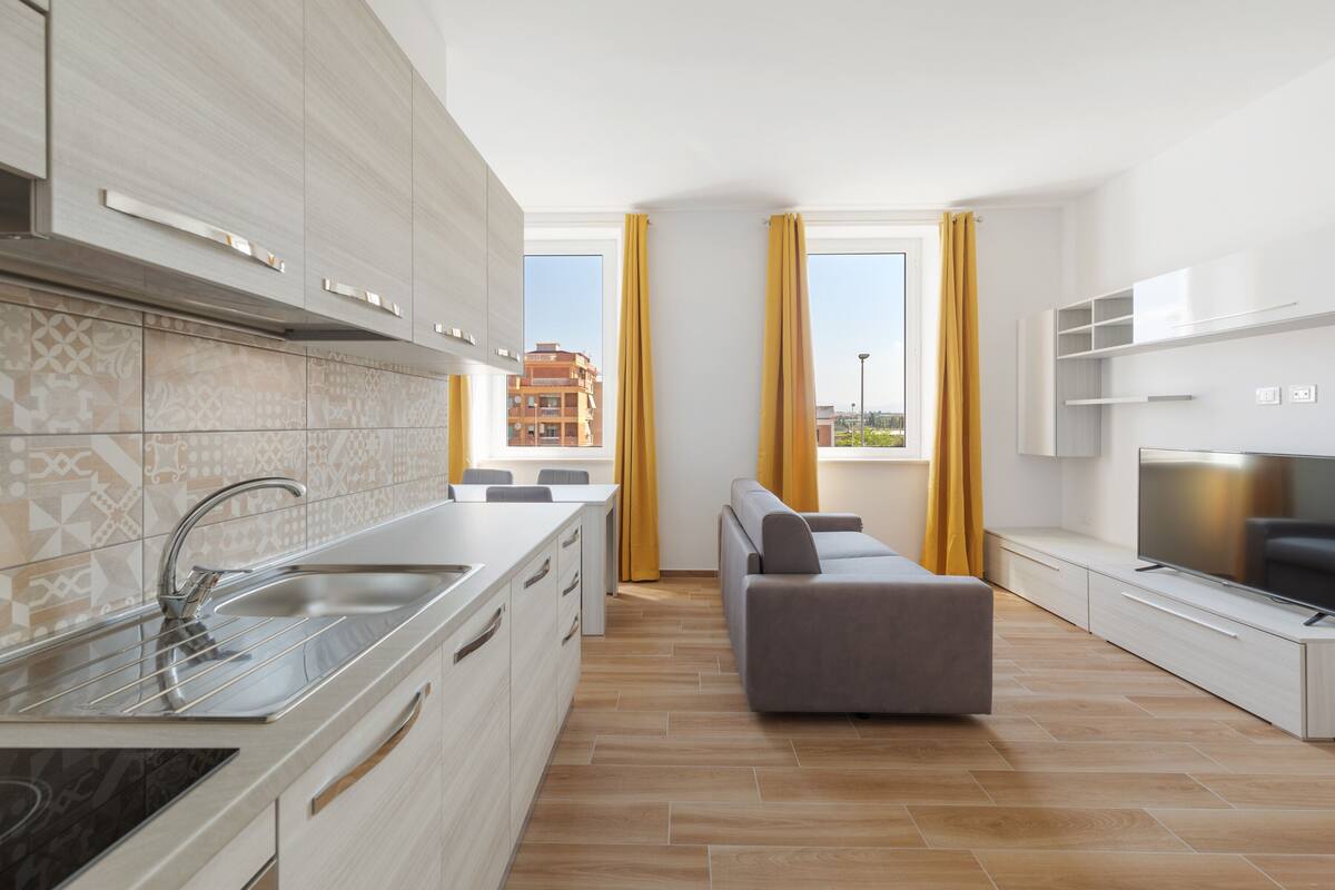 A modern kitchen with a built-in oven, a dining area with a table and chairs, a grey sofa, and a TV on a stand.