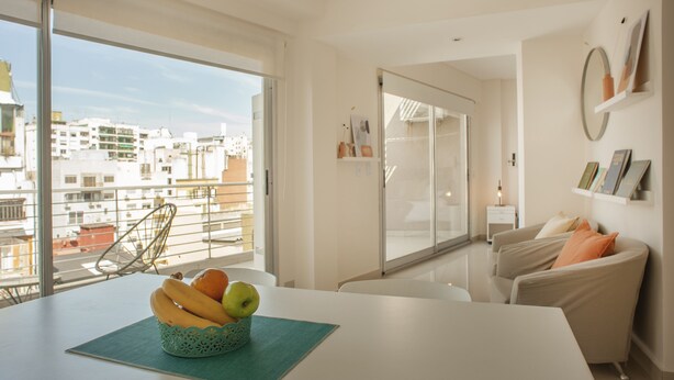 Una sala de estar moderna con balcón, un comedor con frutero y un sofá con cojines.