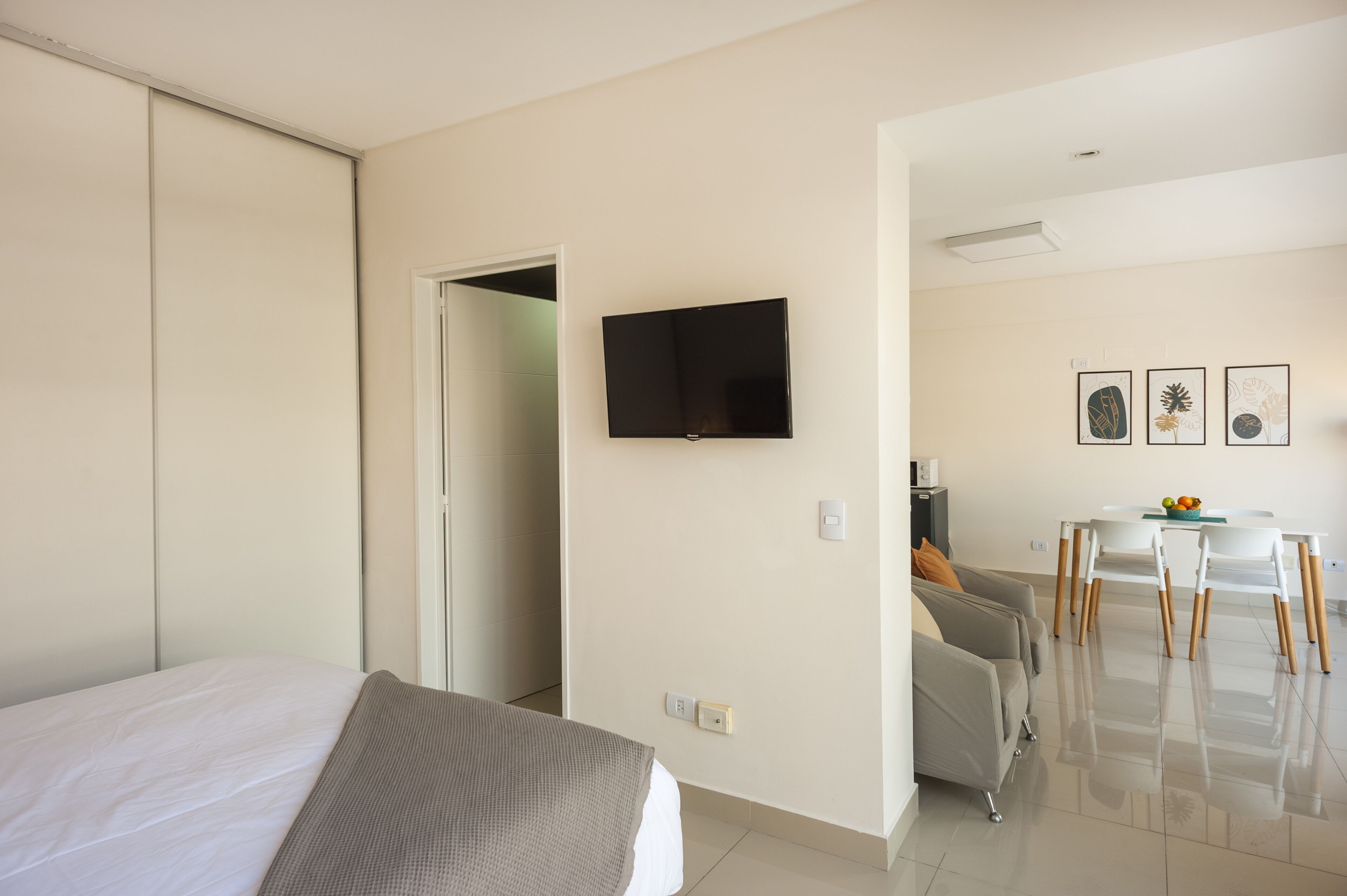 A hotel room with a bed, a TV mounted on the wall, a dining area with a table and chairs, and a kitchenette.