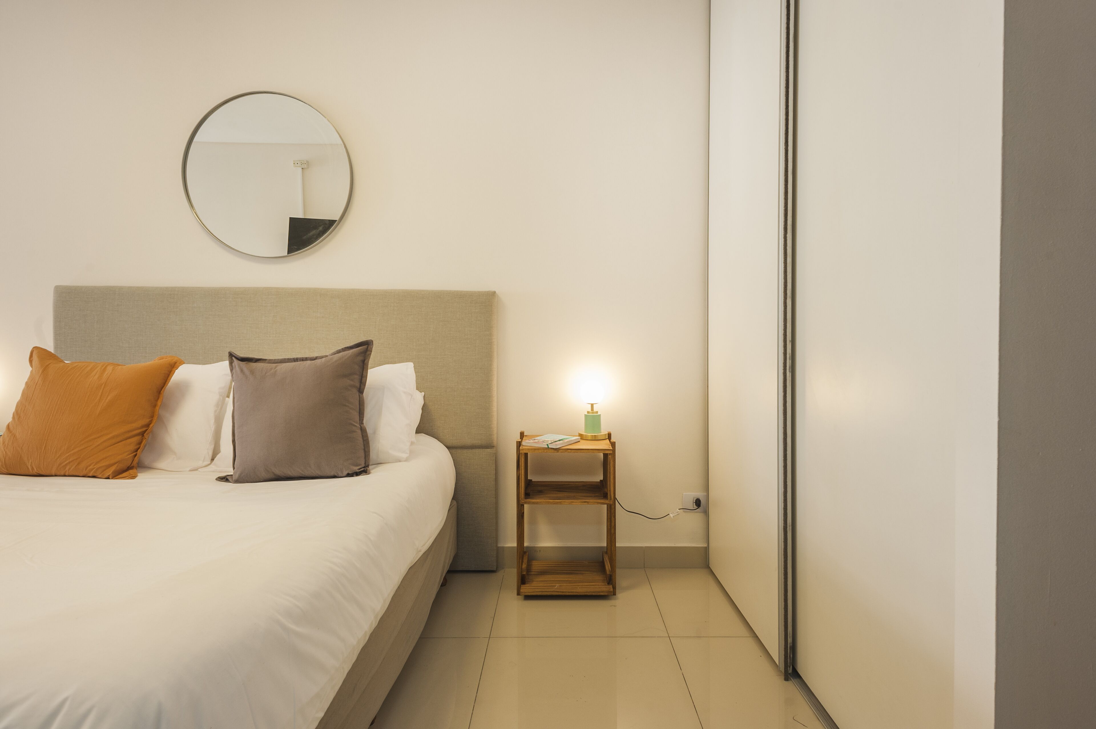 A neatly made bed with a headboard, a bedside table with a lamp, and a round mirror on the wall.