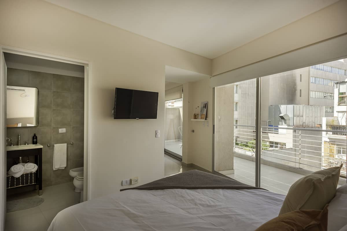 A modern hotel room with a large bed, a bathroom with a shower, a flat-screen TV, and a balcony with a city view.