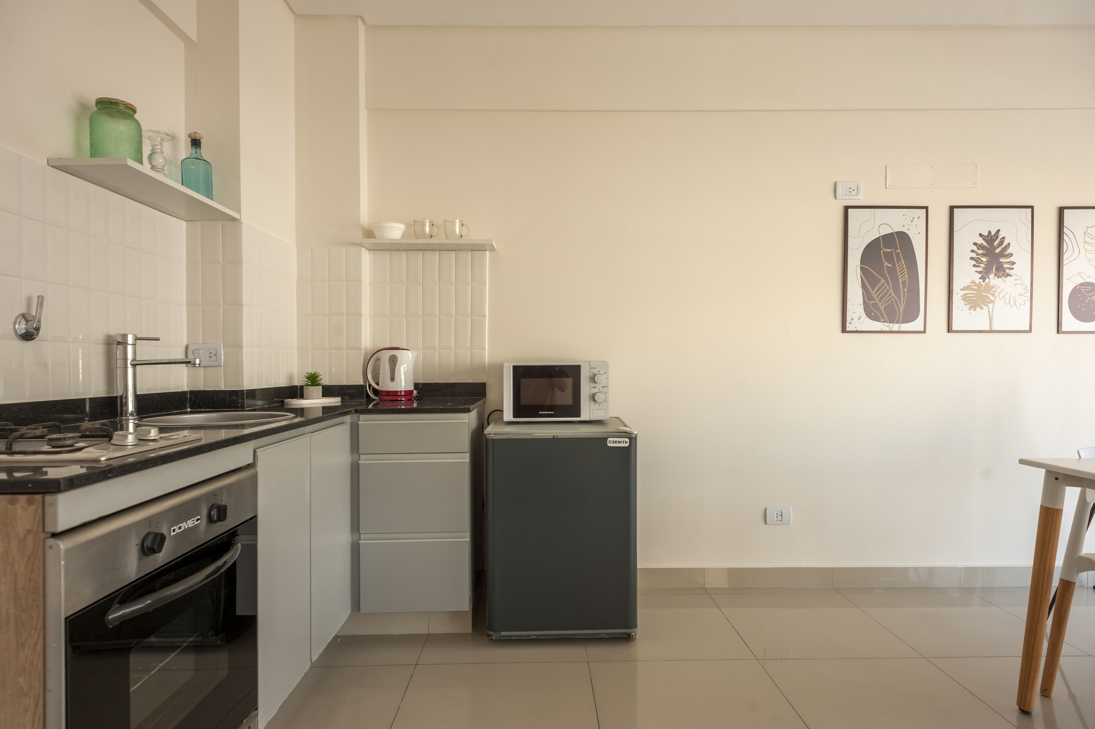 A compact kitchen with a stove, microwave, and small refrigerator. There are shelves with kitchenware and a few framed pictures on the wall.