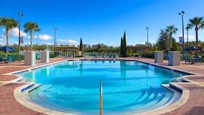 A large, clear swimming pool with a circular design, surrounded by lounge chairs and umbrellas, and palm trees in the background. - Cozy 3 Bedroom Villa Near Disney (Kissimmee)