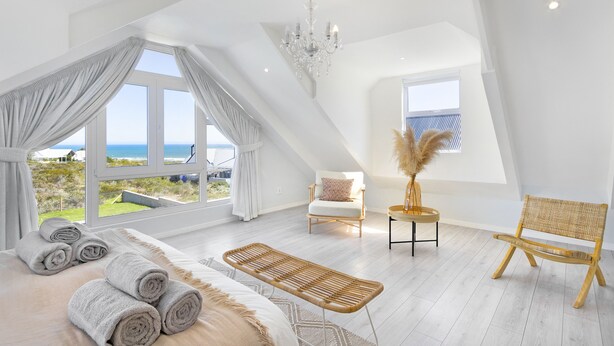 A spacious room with a large window, a bed with rolled towels, a rattan bench, a white chair, and a wooden chair. - Sand Plover 85 (Grotto Bay)