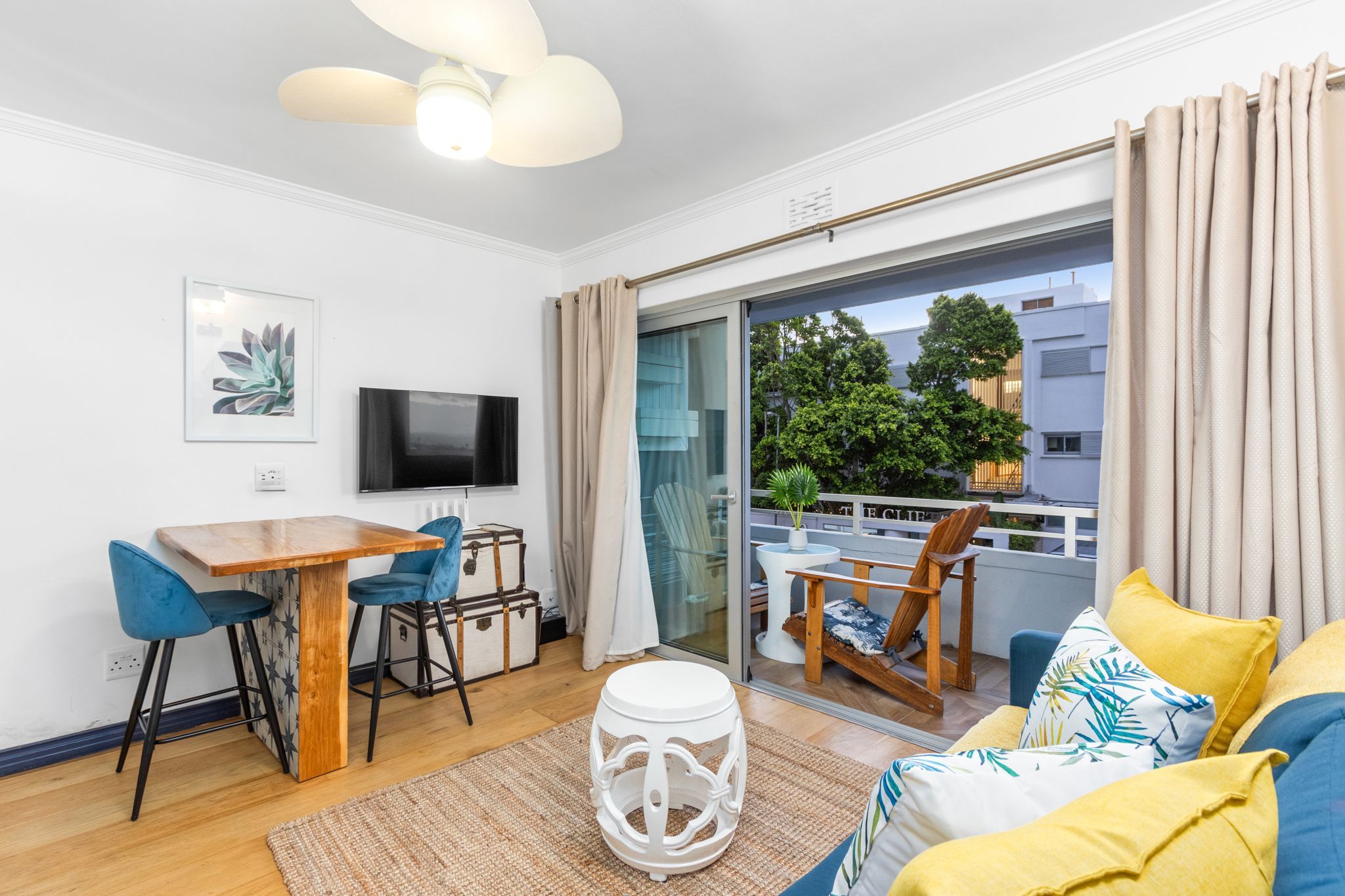 A modern living room with a wooden dining table, blue chairs, a white stool, a flat-screen TV, and a balcony with a view of trees and buildings.
