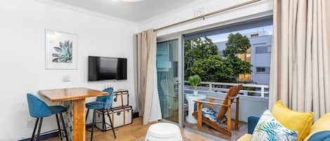 A modern living room with a wooden dining table, blue chairs, a white stool, a flat-screen TV, and a balcony with a view of trees and buildings.