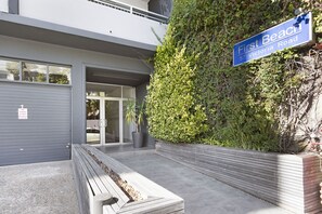 A building with a sign reading "First Beach 57 Victoria Road" and a modern entrance with a bench and planter. - Clifton Studio 104 (Cape Town)