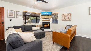 A living room with a fireplace, a ceiling fan, and a large TV. - Outdoor Oasis: HotTub, Fenced, Firepit & PineTrees (Flagstaff)