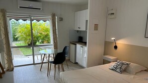 A hotel room with a bed, a desk, a chair, a kitchenette, and a sliding glass door leading to a garden. - Super Appartement Avec Piscine, en Plein Coeur du Centre Ville de la Marina (Le Gosier)