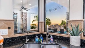 Una cucina con un lavello doppio, una parete decorativa in pietra, un'ampia finestra vista piscina e palme, e una ventola a soffitto.
