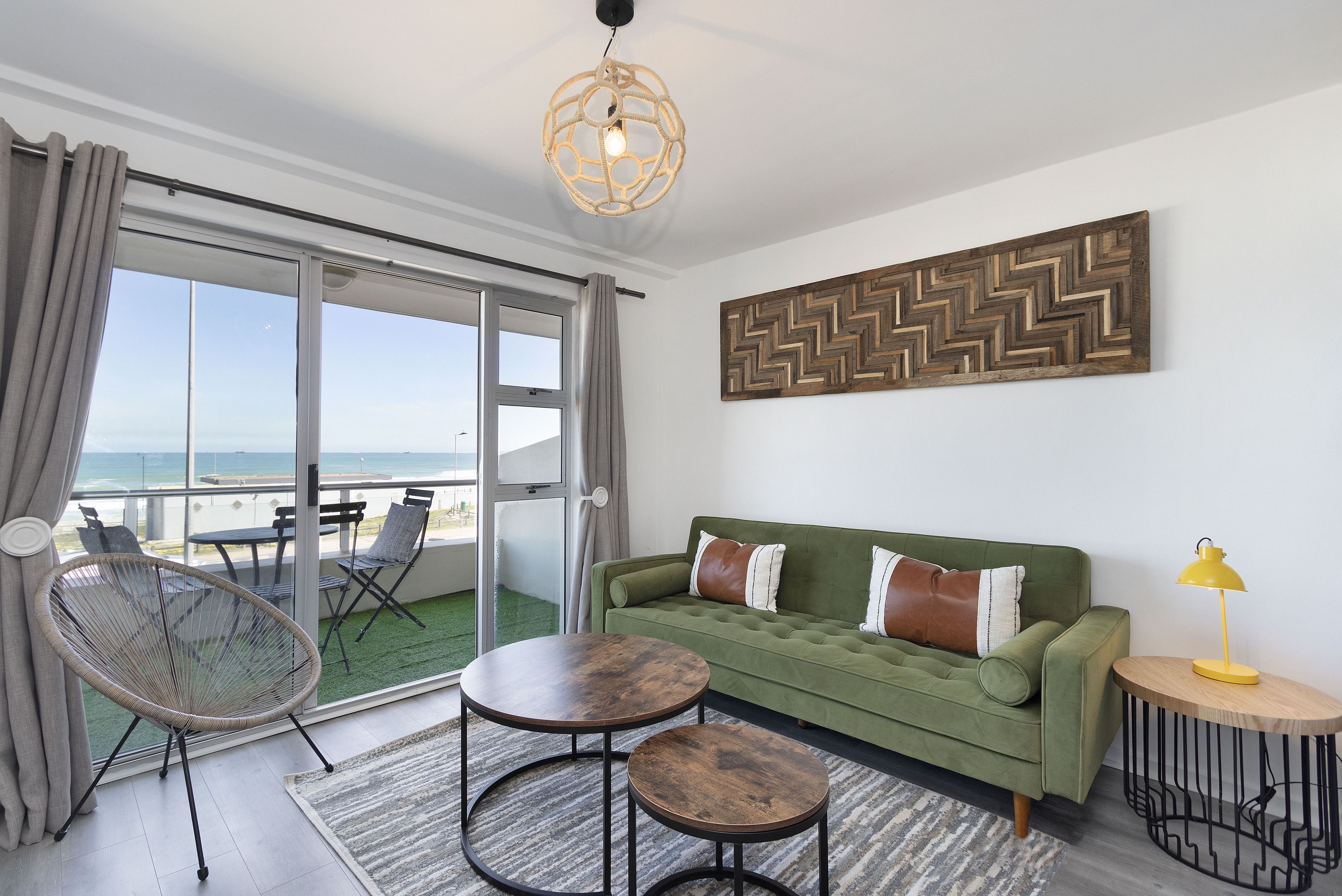 A modern living room with a green sofa, two round tables, a wicker chair, and a view of the ocean through large sliding doors.