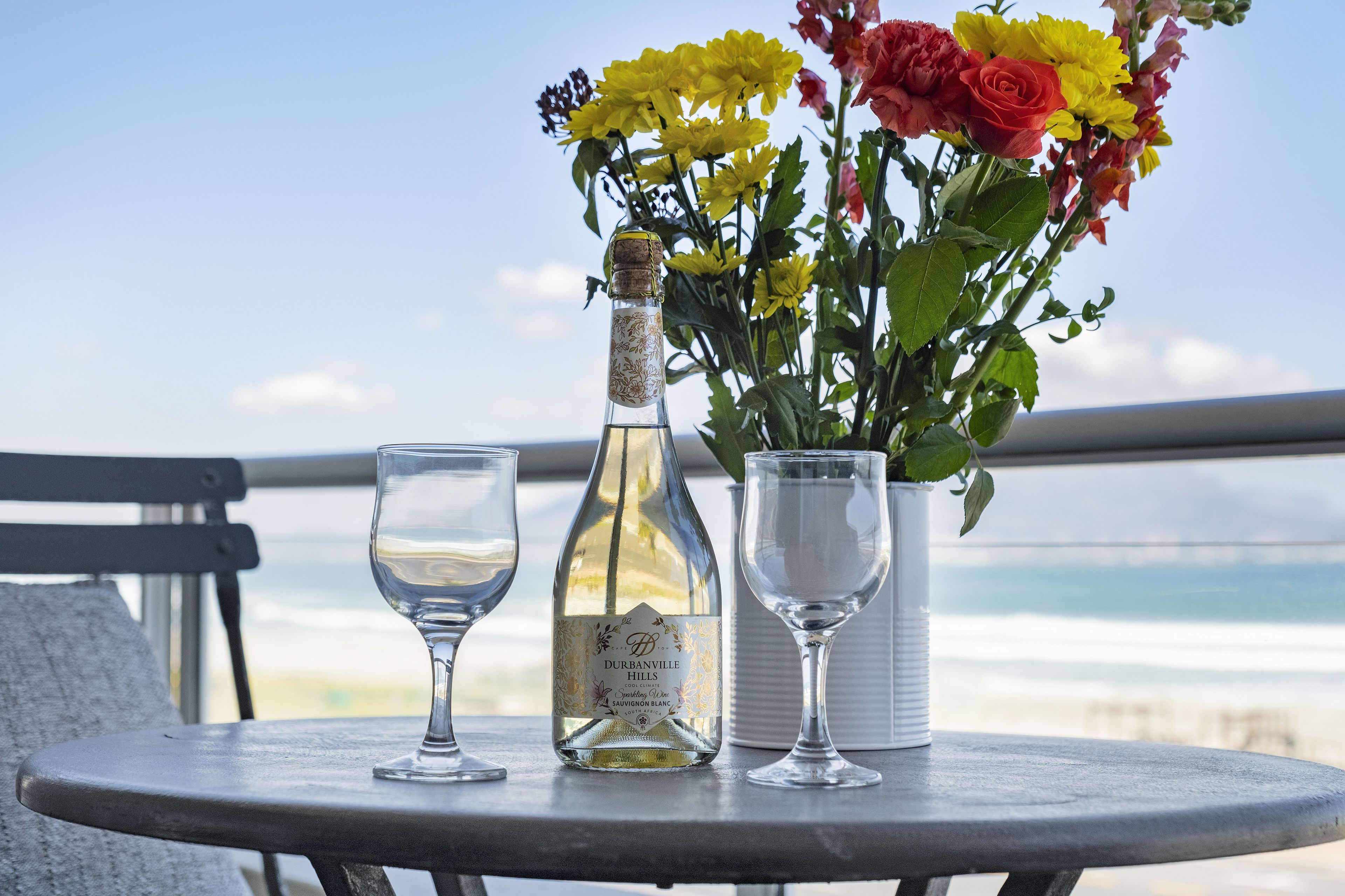 A bottle of Durbanville Hills Sauvignon Blanc, two wine glasses, and a vase with yellow and red flowers on a balcony overlooking the ocean.