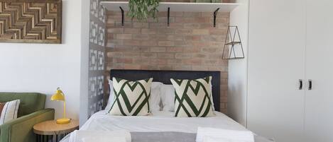 A neatly arranged hotel room with a brick wall, a bed with white linens, green and white patterned pillows, a green armchair, a small round table with a yellow lamp, and a hanging plant.
