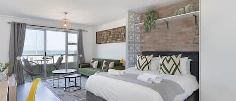 A modern hotel room with a large bed, a green sofa, a dining area, and a view of the sea.