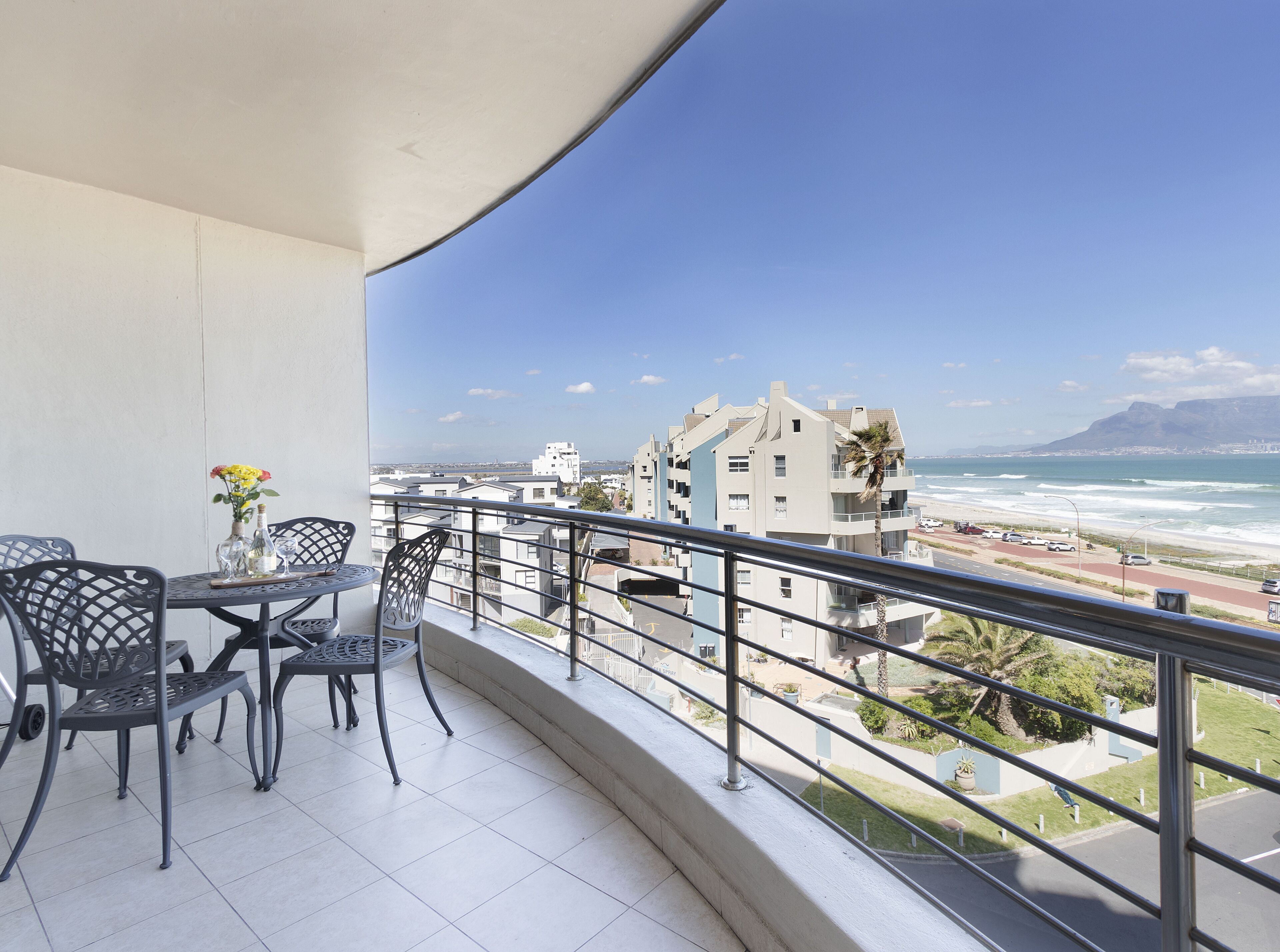 A balcony with a table and chairs, overlooking a coastal view with buildings and a mountain in the distance.