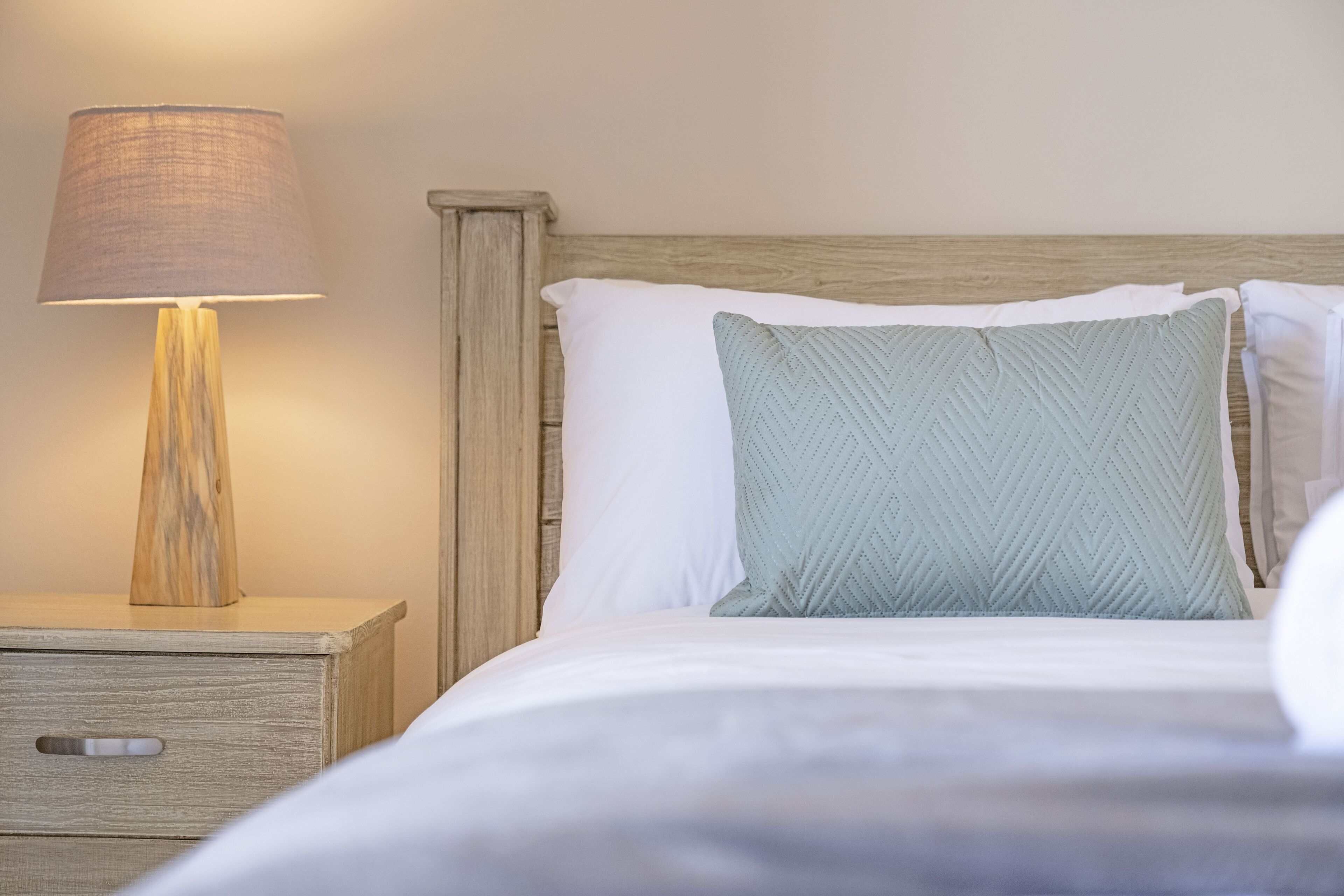 A neatly made bed with a wooden headboard, a bedside lamp, and a small drawer unit.