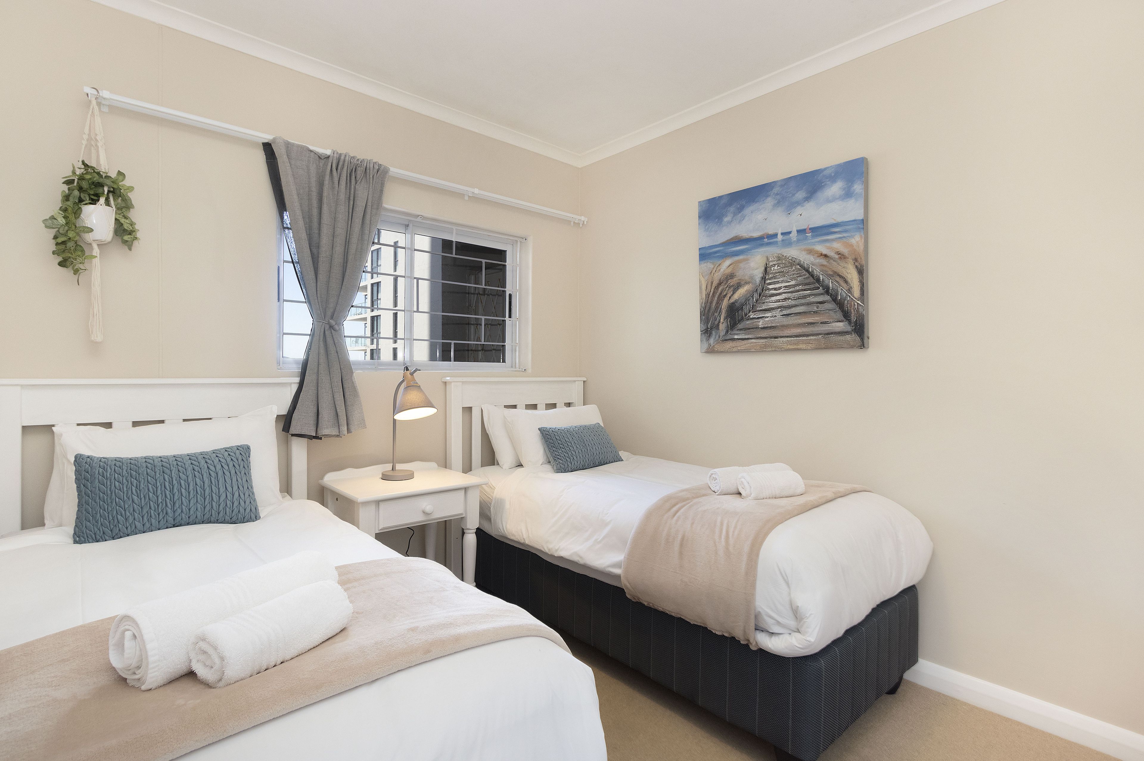 A bedroom with two beds, a window with curtains, a bedside table with a lamp, and a painting on the wall.