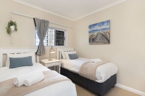 A bedroom with two beds, a window with curtains, a bedside table with a lamp, and a painting on the wall. - Portico 405 (Cape Town)
