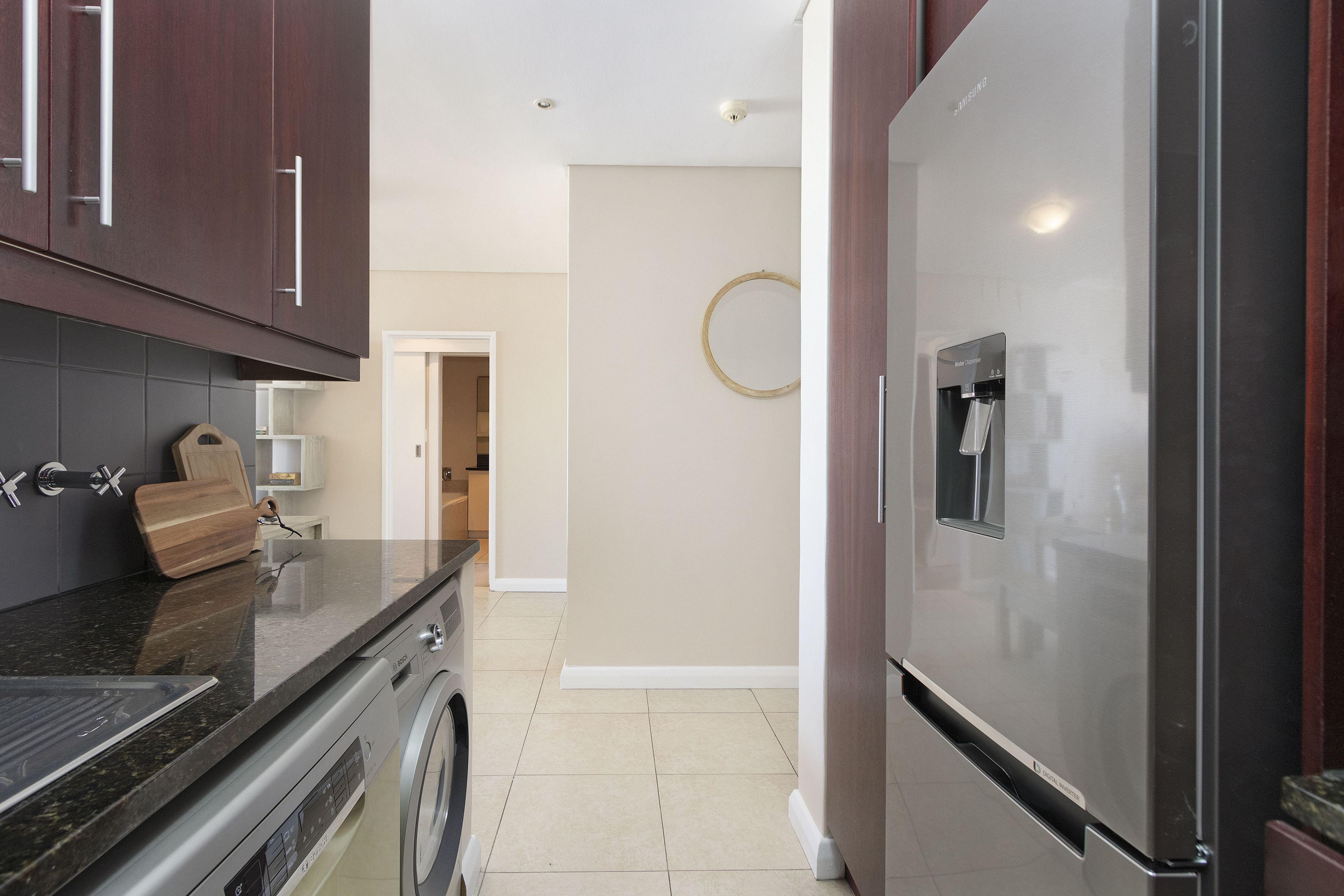 A modern kitchen with dark wooden cabinets, a stainless steel refrigerator, a washing machine, and a countertop with a cutting board.