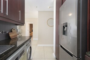 A modern kitchen with dark wooden cabinets, a stainless steel refrigerator, a washing machine, and a countertop with a cutting board. - Portico 405 (Cape Town)
