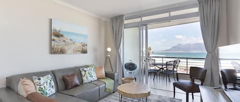 A modern living room with a grey sofa, a round coffee table, and a view of the ocean through large windows.
