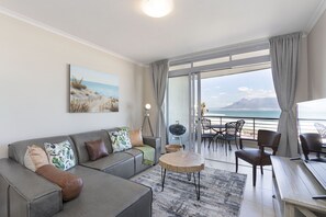 A modern living room with a grey sofa, a round coffee table, and a view of the ocean through large windows. - Portico 405 (Cape Town)