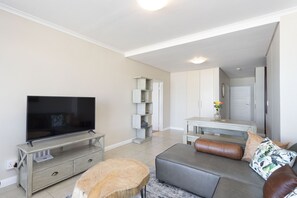 A modern living room with a flat-screen TV, a grey sofa, a wooden coffee table, and a small dining area with a white table and chairs. - Portico 405 (Cape Town)