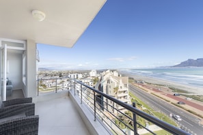 A balcony with wicker furniture, a view of the beach, and a road with cars. - Portico 704 (Cape Town)