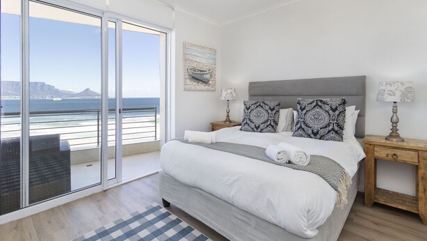 A bedroom with a large bed, a nightstand, a lamp, and a view of the ocean through a sliding door.