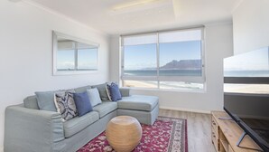 A modern living room with a grey sectional sofa, a large window with a view of the beach, and a flat-screen TV on a wooden stand.