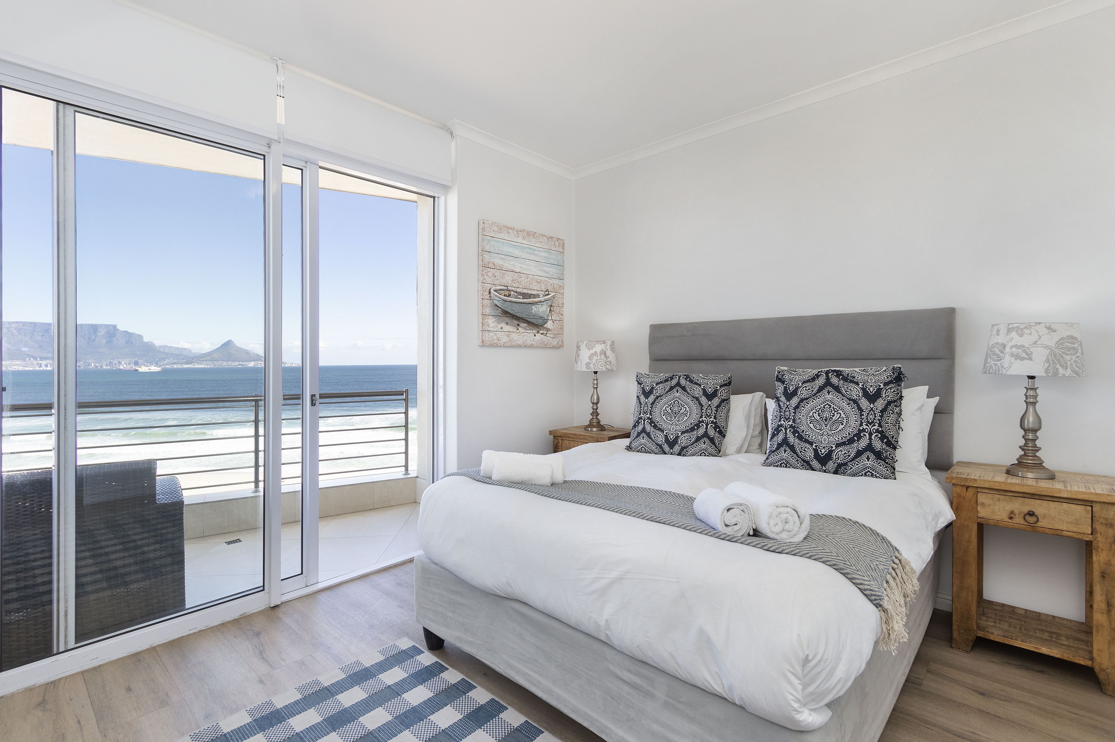 A bedroom with a large bed, a nightstand, a lamp, and a view of the ocean through a sliding door.