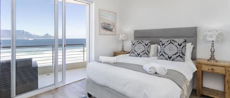 A bedroom with a large bed, a nightstand, a lamp, and a view of the ocean through a sliding door.