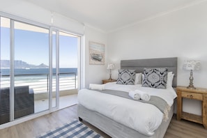 A bedroom with a large bed, a nightstand, a lamp, and a view of the ocean through a sliding door. - Portico 704 (Cape Town)