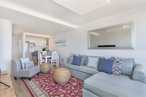A modern living room with a grey sofa, a patterned rug, and a dining area with white furniture.