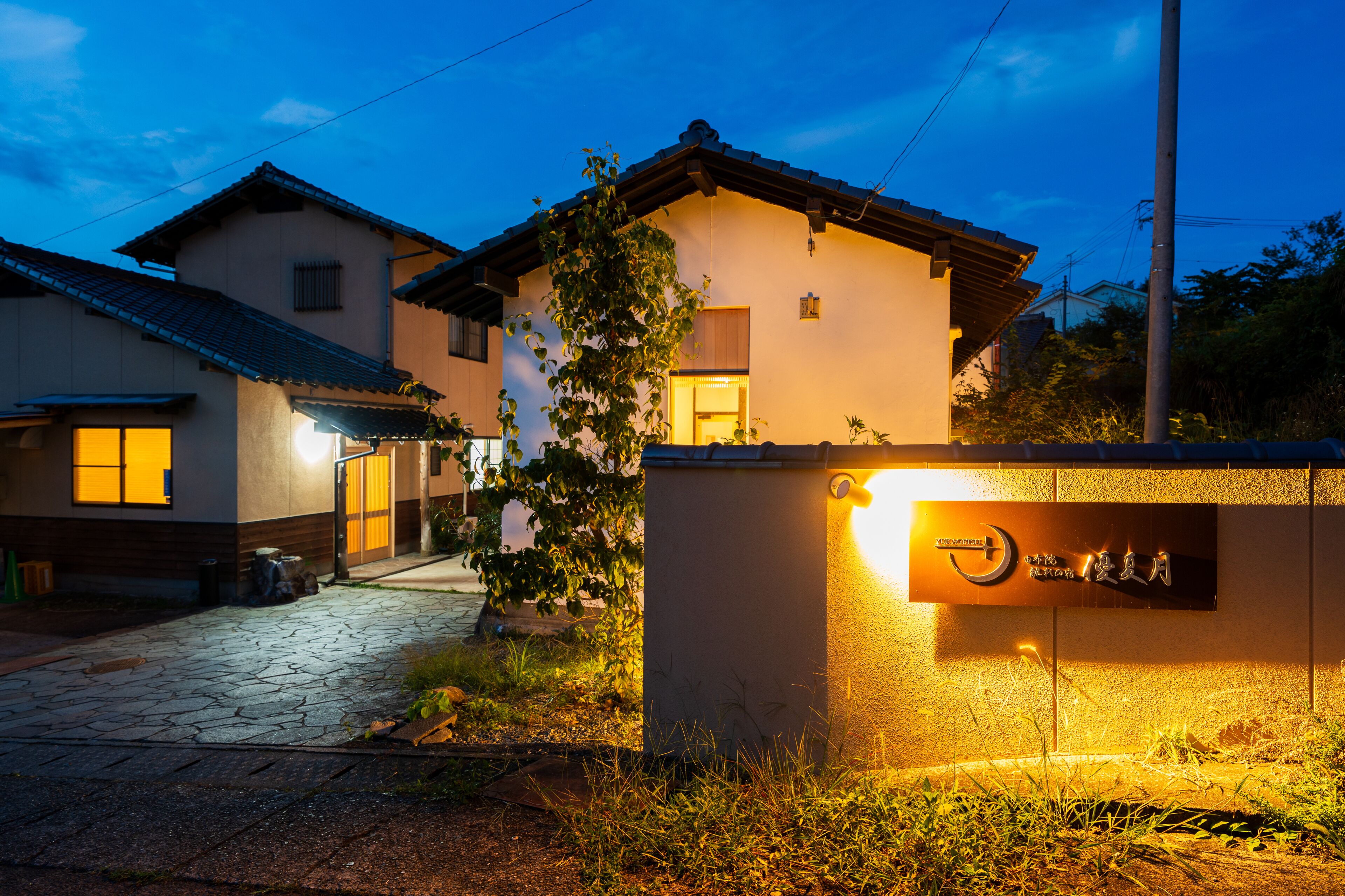 A traditional Japanese house with a stone pathway leading to the entrance, a well-lit signboard, and a clear night sky.