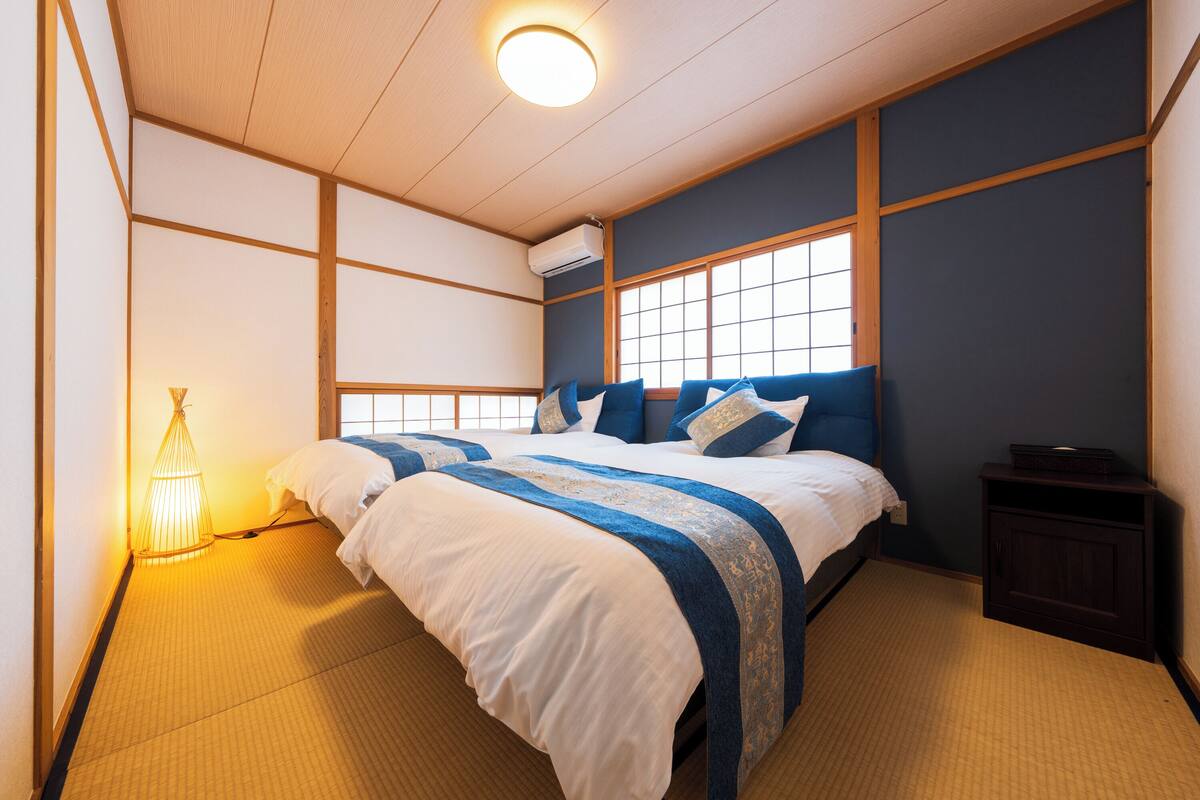 Une chambre de style japonais traditionnel, avec un sol recouvert de tatamis, un lit avec une literie bleue et blanche, une petite table de chevet et une fenêtre avec des cadres en bois.