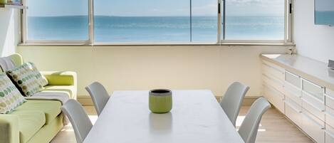 A room with a large window overlooking the sea, a white table with four chairs, a green vase, and a green sofa with patterned pillows.