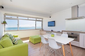 A modern kitchen with a large window, a dining area with a white table and chairs, and a green sofa with a view of the sea. - Sandringham 45 (Cape Town)