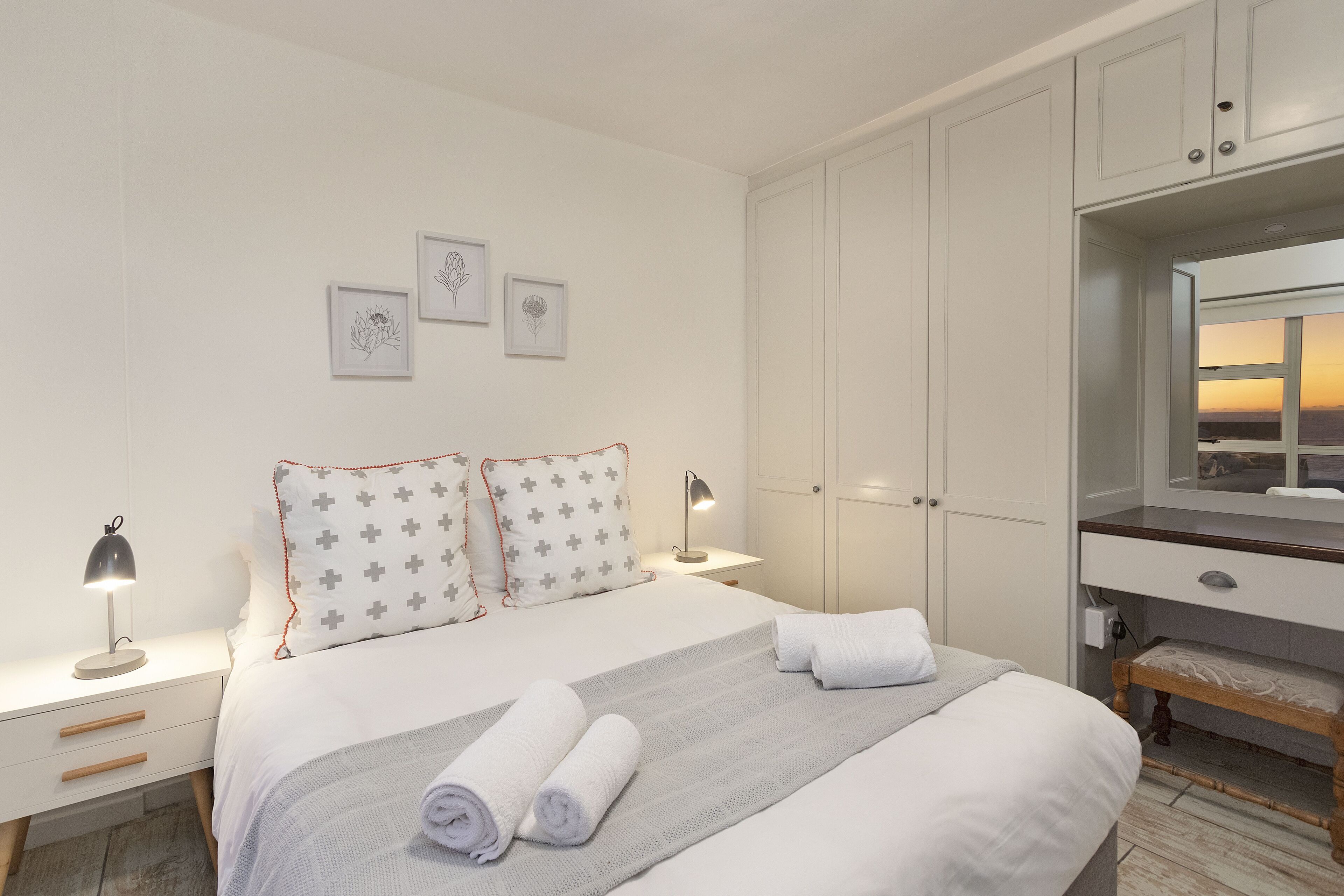 Una cama bien hecha con toallas enrolladas, una lámpara de noche y una mesita de noche. Hay tres cuadros en la pared, un armario y un espejo sobre el tocador.