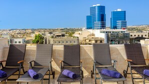A rooftop terrace with four lounge chairs, each with a rolled-up towel. In the background, there are two prominent blue buildings and a cityscape.