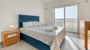 Ein Schlafzimmer mit einem großen Bett, einem Nachttisch, einer Kommode und Blick ins Freie durch eine Schiebetür.