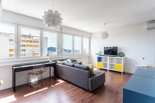 Ein modernes Wohnzimmer mit einer Couch, einem Schreibtisch und einem Fernseher. Große Fenster bieten Blick auf umliegende Gebäude.